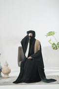 Model wearing a black Kaftan abaya sitting on a chair with a white wall and plant in the background